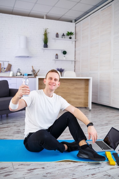 De man gaat thuis sporten. Atleet drinkt water terwijl hij thuis op de grond zit, gezonde levensstijl