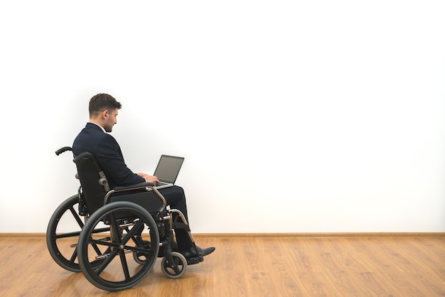 De man in een rolstoel die met laptop op de witte muurachtergrond werkt
