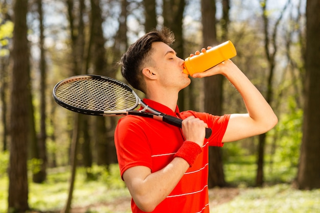 De man in een rood t-shirt poseert met een tennisracket en drinkwater uit een thermokoppel.