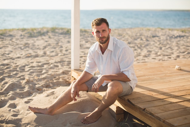 De man in het witte shirt en korte broek zittend op het strand.