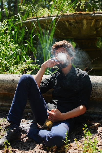 De man met de baard in het donkere shirt en broek zit in een park en rookt een sigaar