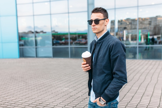 De man met stijlvolle zonnebril, een man loopt door de stad en drinkt koffie uit een papieren beker, een knappe man loopt rond en rust uit, de man op de achtergrond van een modern gebouw, kantoor