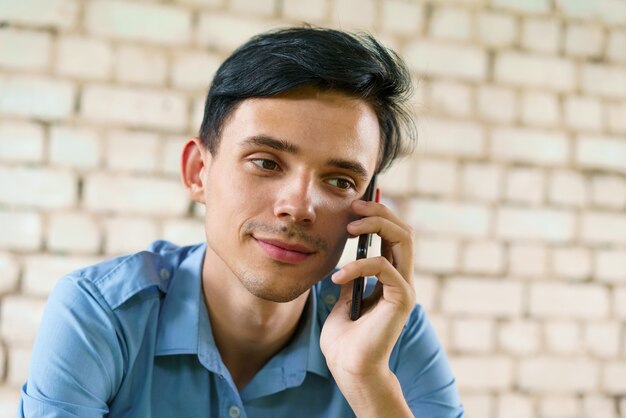 De man praat aan de telefoon close-up Kaukasische jonge brunette man op witte bakstenen muur achtergrond...