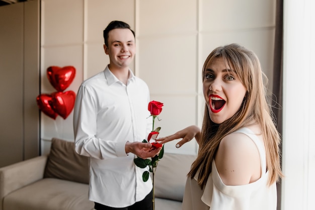 De man stelt aan vrouw met een ring en rode roos thuis met hartvormige ballonnen voor