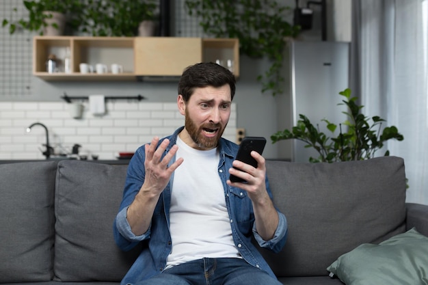 De man zelf thuis geschokt en teleurgesteld kijkt naar de telefoon zittend op de bank