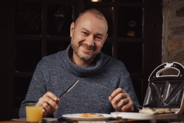 De man zit te ontbijten Een man van middelbare leeftijd in een trui bereidt zich voor om een broodje te eten in een donkere kamer