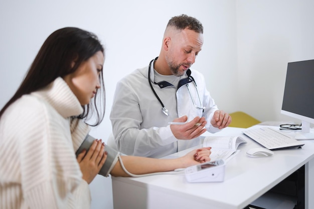 De mannelijke arts meet de bloeddruk van de vrouw en noteert de resultaten op de kaart van de patiënt. Vrouw bij de afspraak bij cardioloog