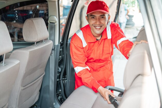 De mannelijke autoreiniger draagt een glimlachend rood uniform tijdens het schoonmaken van de stofzuigerstoel in de auto