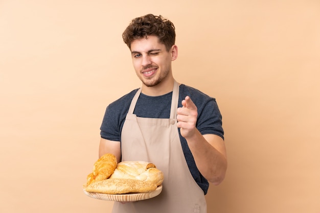 De mannelijke bakker die een lijst met verscheidene die brood houden op beige muur wordt geïsoleerd richt vinger op u