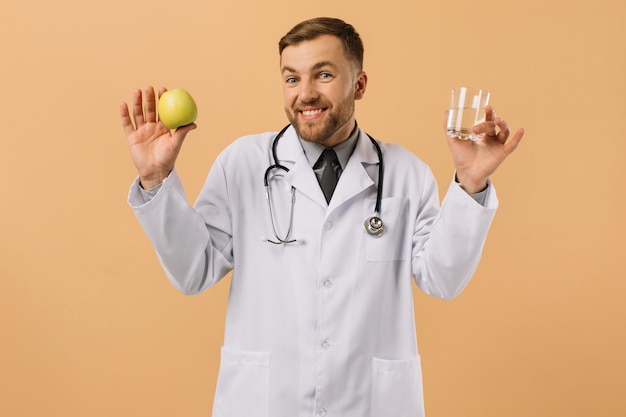 De mannelijke voedingsdeskundige arts met een stethoscoop glimlachend en vasthoudend water en appel op beige achtergrond dieetplan concept
