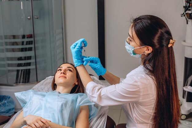 De medewerker van de schoonheidssalon bereidt de spuit voor door deze te vullen met Botox om tekenen van veroudering op het gezicht van een meisje dat op de bank naast haar ligt te voorkomen.