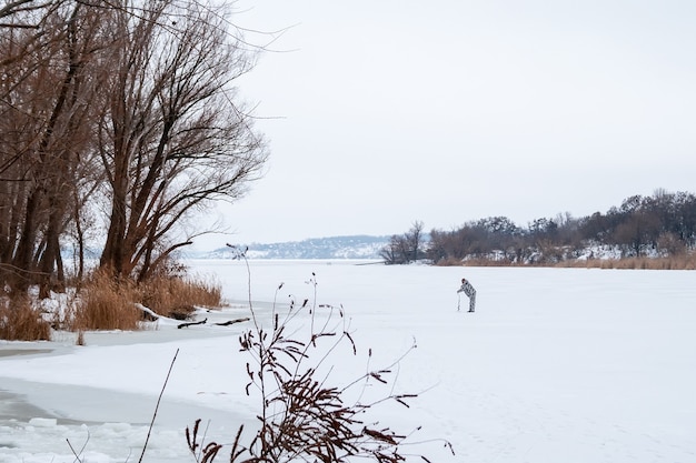 De mens boort een gat om te vissen op een besneeuwde bevroren rivier in de verte. Winterlandschap