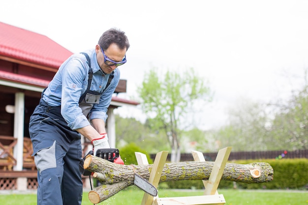 De mens in overall zaagt hout met kettingzaag gebruikend zaagbok