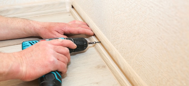 De mens installeert de plint met een boor. Reparatie werkt binnen. Renovatie in de flat.