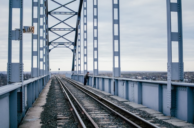 De mens staat verveeld op rails van een oude spoorbrug, helaas wachtend op een vergadering. Industrieel landschap. Grote ijzeren brug op een bewolkte dag. Het concept van scheiding, ontmoeting, depressie. Kopieer ruimte