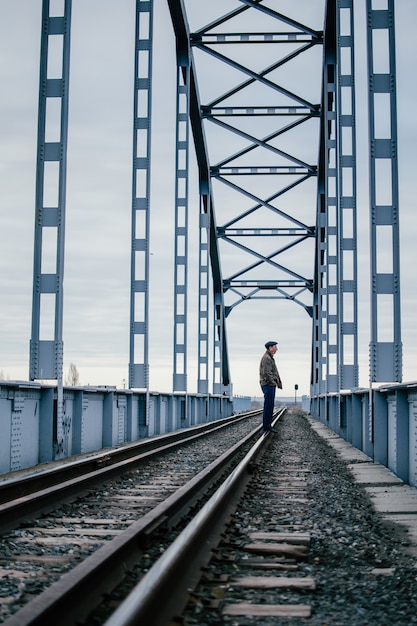 De mens staat verveeld op rails van een oude spoorbrug, helaas wachtend op een vergadering. Industrieel landschap. Grote ijzeren brug op een bewolkte dag. Het concept van scheiding, ontmoeting, depressie. Kopieer ruimte