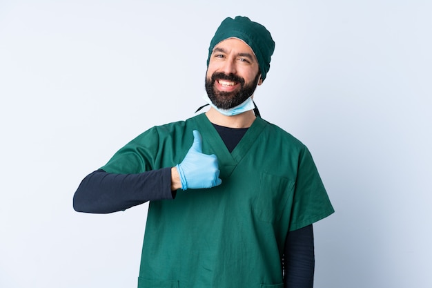 De mens van de chirurg in groen uniform over geïsoleerde muur die een duim omhoog gebaar geeft