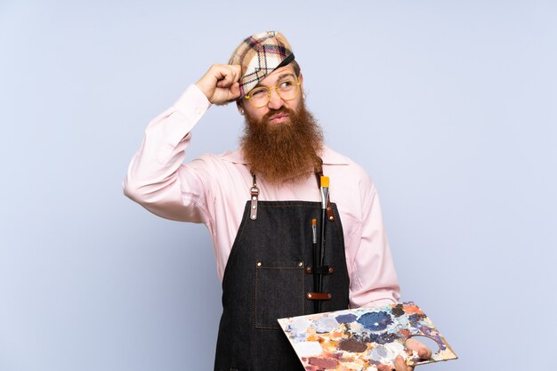 De mens van de roodharigekunstenaar met lange baard die een palet over geïsoleerde blauwe muur houden die twijfels hebben en met verwarren gezichtsuitdrukking