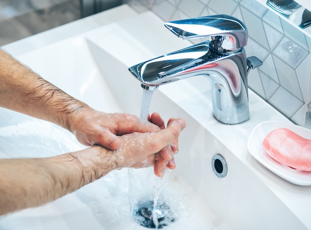 De mens wast handen met zeep en warm water bij de gootsteen van de huisbadkamer. Guy maakt handen schoon. Hygiëne voor preventie van coronavirusuitbraak. Covid-19 pandemische bescherming door regelmatig de handen te wassen