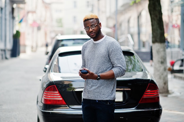 De modieuze mens op grijze sweater en glazen stelde op straat tegen zwarte bedrijfsauto en sprekend op telefoon