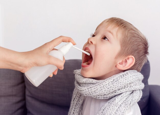 De moeder van een kleine jongen gebruikt een helende spray voor zijn keel. Ziek kind bevriest verpakt in een sjaal. Geneesmiddel tegen hoest.