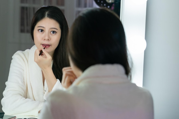 De mooie Aziatische make-up van de vrouwenkunstenaar paste rode lippenstift toe door borstel. Japans jong schoonheidsmodelmeisje die badjas schilderende lippen dragen die spiegel in kleedkamer backstage bekijken. zangeres maak je klaar.