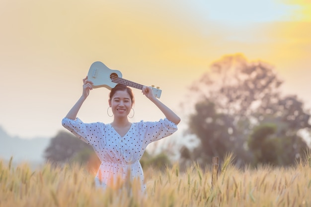 De mooie Aziatische ukelele van de vrouwenholding en het ontspannen op gerstgebied in de zomer op zonsondergangtijd