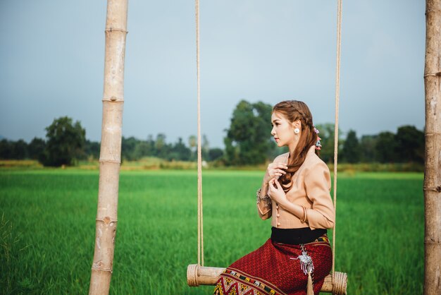 De mooie aziatische vrouw in lokale kledingszitting op schommeling en geniet van natuurlijk op bamboebrug in padieveld