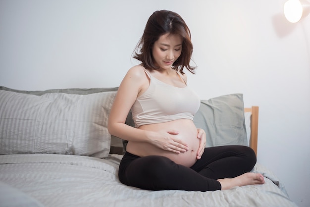 De mooie Aziatische zwangere vrouw zit op bed en wat betreft haar buik