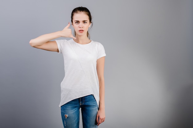 De mooie donkerbruine vrouw die het witte overhemd tonen dragen roept me gebaar die telefoon met hand imiteren die over grijs wordt geïsoleerd