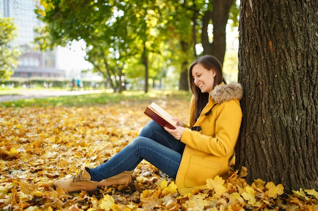 De mooie gelukkig lachende bruinharige vrouw in gele jas en spijkerbroek zittend onder de esdoorn met een rood boek in herfst stadspark op een warme dag. Herfst gouden bladeren. Leesconcept