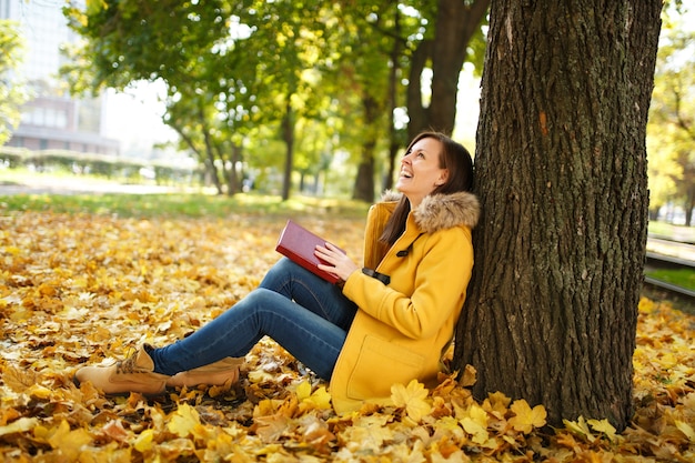 De mooie gelukkig lachende bruinharige vrouw in gele jas en spijkerbroek zittend onder de esdoorn met een rood boek in herfst stadspark op een warme dag. Herfst gouden bladeren. Leesconcept