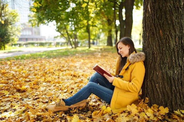 De mooie gelukkig lachende bruinharige vrouw in gele jas en spijkerbroek zittend onder de esdoorn met een rood boek in herfst stadspark op een warme dag. Herfst gouden bladeren. Leesconcept