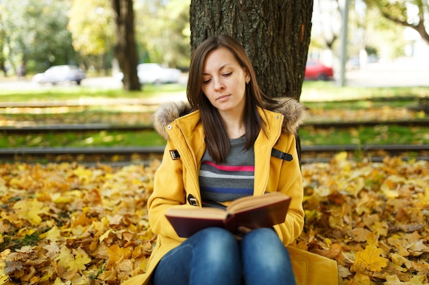 De mooie gelukkig lachende bruinharige vrouw in gele jas en spijkerbroek zittend onder de esdoorn met een rood boek in herfst stadspark op een warme dag. Herfst gouden bladeren. Leesconcept