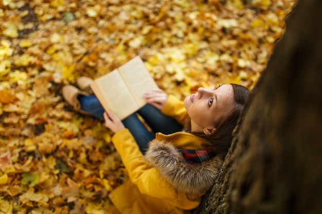 De mooie gelukkig lachende bruinharige vrouw in gele jas en spijkerbroek zittend onder de esdoorn met een rood boek in herfst stadspark op een warme dag. Herfst gouden bladeren. Lezingsconcept. Bovenaanzicht.