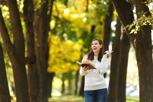 De mooie gelukkig lachende bruinharige vrouw in witte trui staande met een rood boek in herfst stadspark op een warme dag. Herfst gouden bladeren. Lezingsconcept.