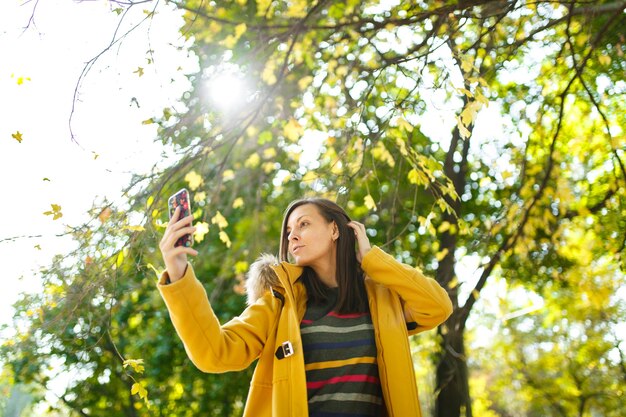 De mooie gelukkige vrolijke bruinharige vrouw in een gele jas en gestreepte longsleeve doet selfie op mobiele telefoon in herfst stadspark op een warme dag. Herfst gouden bladeren.