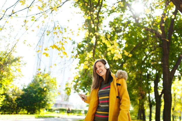 De mooie gelukkige vrolijke bruinharige vrouw in een gele jas en gestreepte longsleeve verheugt zich met een tablet in haar handen en witte koptelefoon in herfst stadspark op een warme dag. Herfst gouden bladeren.