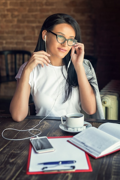 De mooie glimlachende vrouw in glazen luistert muziek door haar smartphone bij koffie.