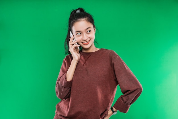 De mooie jonge Aziatische vrouw spreekt aan de telefoon die vaag glimlacht met iemand terwijl zij naast geïsoleerd tegen de groene achtergrond kijkt