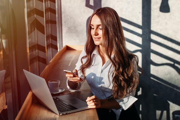 De mooie jonge bedrijfsvrouw in witte blouse die laptop en smartphone met behulp van, drinkt koffie bij een lijst in een koffie