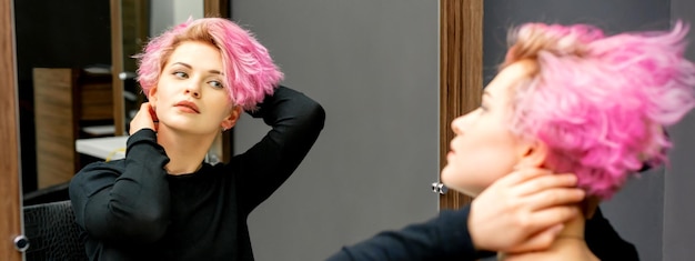 De mooie jonge blanke vrouw met een nieuw kort roze kapsel kijkt naar haar spiegelbeeld in de spiegel en controleert haar kapsel in een kapsalon