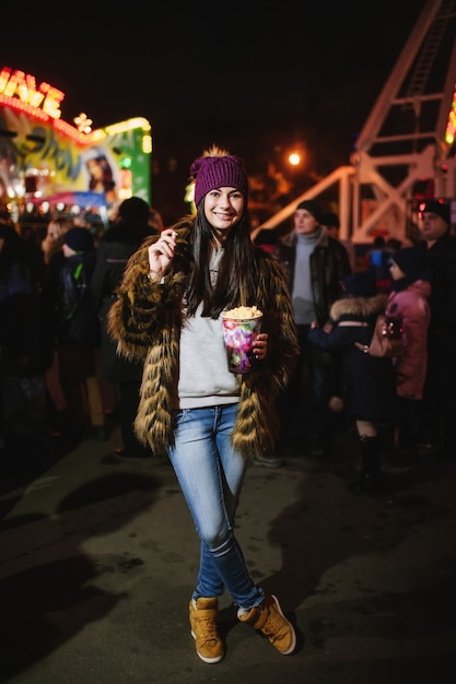 De mooie jonge dame die op de kerstmarkt in de buurt van de carrousel staat en popcorn eet