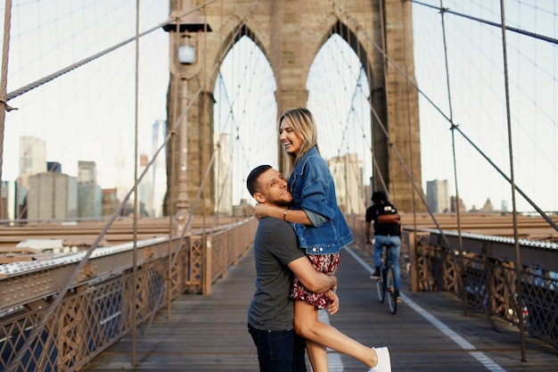 De mooie jonge man en de vrouw stellen op de Brug van Brooklyn in de stralen van ochtendzon