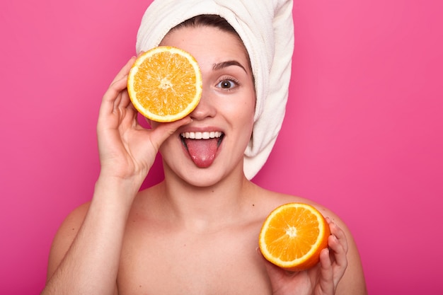 De mooie jonge vrouw houdt de helft van sinaasappel in haar hand en tegens oog met een ander deel van fruit. aantrekkelijke dame met handdoek op hoofd staat op roze. dagelijkse procedure en schoonheidsconcept.
