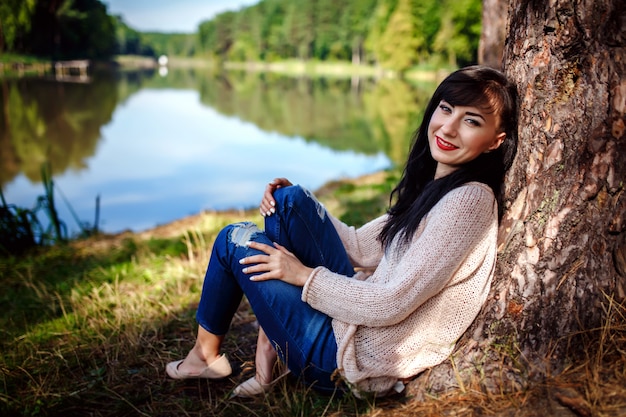 De mooie jonge vrouw in jeans zit op rivieroever dichtbij grote boom in zonnige dag