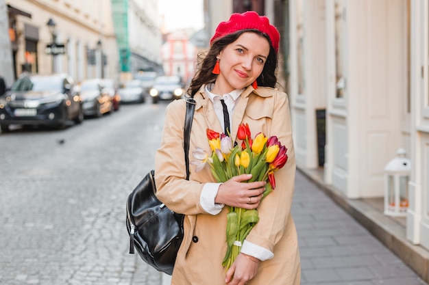 De mooie jonge vrouw met de bloemenboeket van de lentetulpen bij stadsstraat. Gelukkig meisje buiten lopen. Lente portret van mooie vrouw in de oude stad