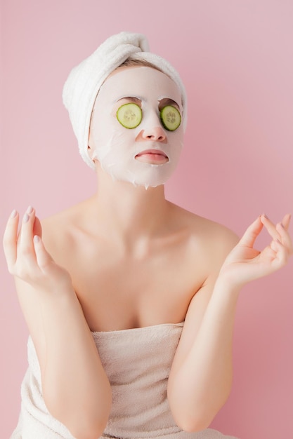 De mooie jonge vrouw past een kosmetisch weefselmasker op een gezicht met komkommer op een roze achtergrond toe.
