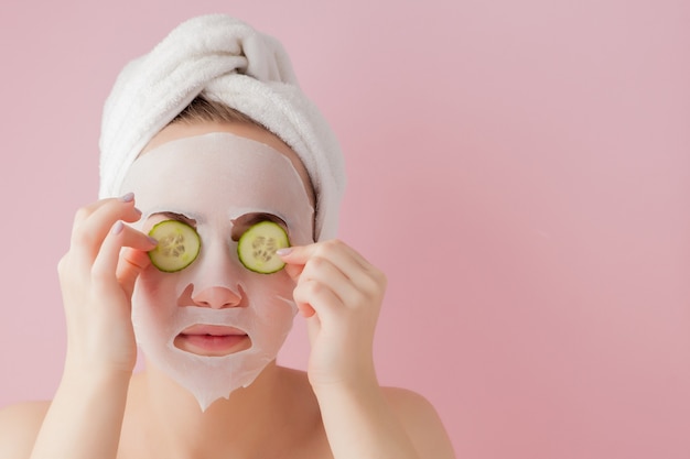 De mooie jonge vrouw past een kosmetisch weefselmasker toe op een gezicht met komkommer op een roze ruimte