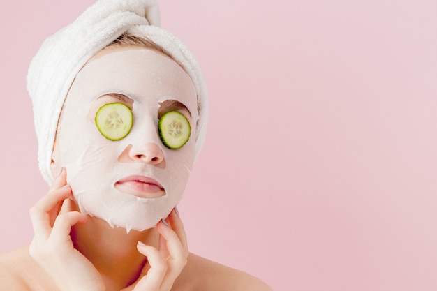 De mooie jonge vrouw past een kosmetisch weefselmasker toe op een gezicht met komkommer op roze.